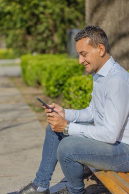 Genç Latin bir adam kredi kartıyla alışveriş yapıyor. Cep telefonuyla bankta oturuyor..