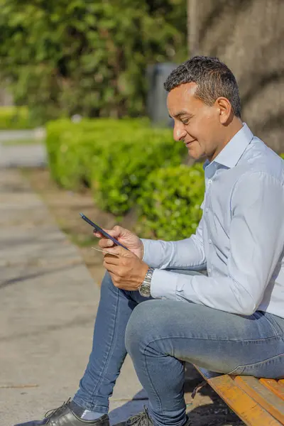 Young Latino man making a purchase with a credit card on his mobile phone sitting on a bench.