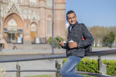 Elinde tek kullanımlık kahve tutan Latin bir adam halka açık bir parkta üzerinde fotokopi alanı olan bir tırabzana yaslanıyor..