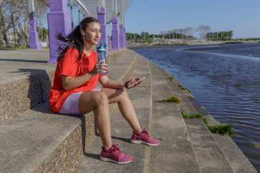 Spor kıyafetli bir kız merdivenlerde oturmuş bir şişe sudan içiyor..