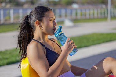 Spor giyinen Latin bir kız dışarıda egzersiz makinesinde su içiyor..