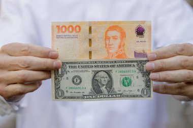 Bin Arjantin pesosunun yanında bir dolarlık banknot tutan bir adamın yakın çekimi..