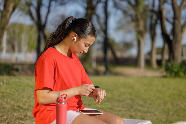 Latin girl with wireless headphones sitting on a bench in a public park checking her smart watch.