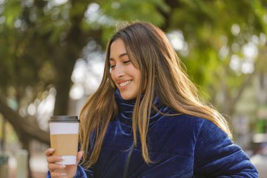 Gülen Latin kız halka açık bir bankta kahve içiyor..