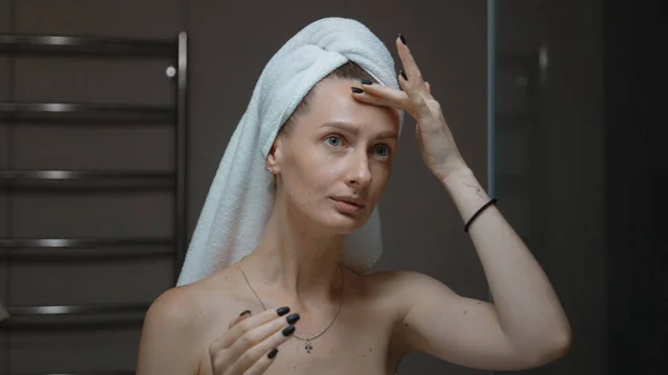 Woman with towel wrapped around her head massaging her face before bath mirror. Daily evening routine - facial cleaning, skin care, peeling beauty treatment concept