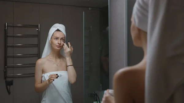 Woman with towel wrapped around her head massaging her face before bath mirror. Daily evening routine - facial cleaning, skin care, peeling beauty treatment concept