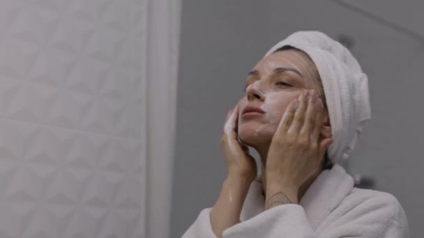 タオルを頭に巻きつけた若い女性が洗顔フォームで顔の肌を洗います 毎日の朝のルーチン 顔のクリーニング スキンケア 保湿と美容トリートメントのコンセプト — ストック動画