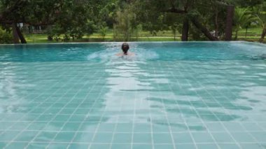A beautiful long-haired woman swims and sits on the edge in an outdoor infinity pool against the backdrop of tropical vegetation. High quality 4k footage