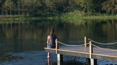 A woman in a blouse sits on a wooden pier by the lake in summer or autumn with bright trees and reflections in the water. High quality 4k footage