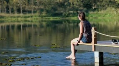 A woman in a blouse sits on a wooden pier by the lake in summer or autumn with bright trees and reflections in the water. High quality 4k footage