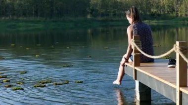 A woman in a blouse sits on a wooden pier by the lake in summer or autumn with bright trees and reflections in the water. High quality 4k footage