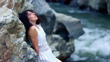 Zarif giyinmiş sakin bir kadın kayalık bir zemine yaslanır, düşünceli bir şekilde akan suya bakar, doğanın güzelliği arasında bir huzur anı yakalar. Yüksek kalite 4k görüntü
