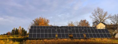Bulutları ve sarı sonbahar ağaçlarıyla dolu mavi gökyüzünün arka planında fotoelektrik bataryalar. Güneş panelleri. Alternatif enerji kavramı