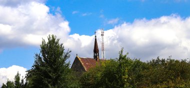 Uzun, eski bir Gotik Katolik kilisesinin kulesi ile telekomünikasyon kulesinin birleşimi mavi gökyüzüne karşı bulutlar. Din ve Katoliklik. Etrafta ağaçlar ve yeşillikler var..