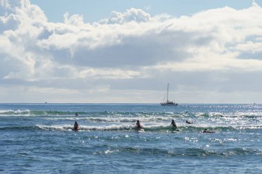 Denizde sörf yapmaktan hoşlanan bir grup birey uzak mesafeden görülebilen bir yelkenliyle resim gibi ufukta ve bulutlu bir gökyüzünde