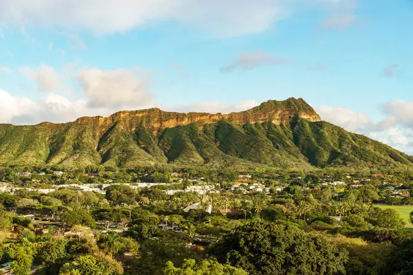 Bir hava manzarası, ağaçlar ve evlerle çevrili bir dağı yakalar, bulutlar, gökyüzü ve çimenler gibi elementlerle resmedilmiş doğal bir manzara oluşturur.