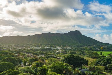 Hava manzarası ağaçlar ve evler tarafından kucaklanan bir dağı yakalıyor, çarpıcı bir doğal manzaranın zeminine kurulmuş ve gökyüzünde bulutlar sürükleniyor.