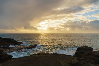 Görüntü, önünde kayalar olan bir su kütlesinin üzerinde sakin ve doğal bir manzara gösteren güzel bir günbatımını tasvir ediyor.