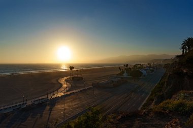 Güneş güzel kumlu bir plajın üzerinde batarken, palmiye ağaçları ön planda yavaşça sallanarak, Santa Monica 'da güzel ve huzurlu bir manzara yaratıyor.