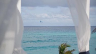 Uçurtma sörfçülerinin zarif bir şekilde uzaklarda süzüldüğü, perdelerle çerçevelenmiş okyanus manzarası.