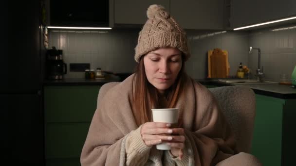 年轻女人戴着用格子花包起来的帽子 一个人坐在没有暖气的公寓里 在寒冷中发抖 没有中央暖气 不健康的女性感到不适 尽量在室内热身 — 图库视频影像