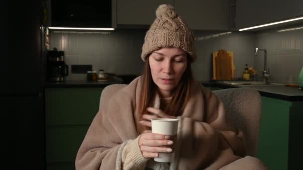 年轻女人戴着用格子花包起来的帽子 一个人坐在没有暖气的公寓里 在寒冷中发抖 没有中央暖气 不健康的女性感到不适 尽量在室内热身 — 图库视频影像