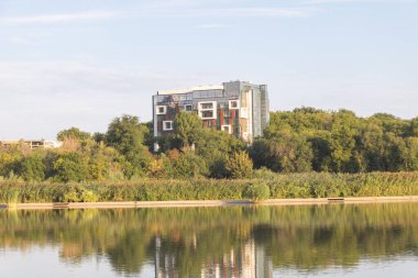 A modern apartment building with glass and metal facades stands on the bank of an industrial lake surrounded by trees and greenery. clipart