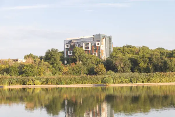 stock image A modern apartment building with glass and metal facades stands on the bank of an industrial lake surrounded by trees and greenery.