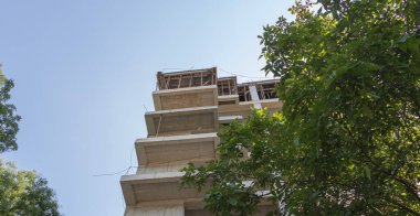 Beyaz beton kaplamalı yüksek bir bina. En üst katlar inşaat halinde, sokak seviyesinden görülüyor..