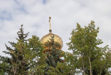 Tepesinde Ortodoks haçı olan altın kubbenin fotoğrafı, büyük bir şehrin ortasında ağaçların arasından görülüyor..