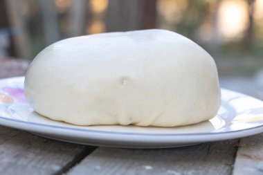 Tabakta büyük yuvarlak bir hamur parçası, yuvarlak şekilli ve pürüzsüz desenli beyaz ve yumuşak görünümlü bir krem peynir..