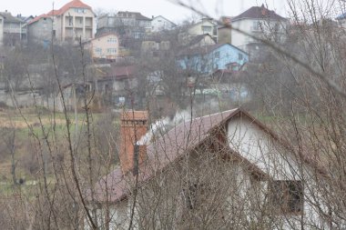 Ön planda, bacası duman çıkaran bir ev - Chisinau 'daki küçük evleri tepeden gören, yaprakları olmayan binaları ve ağaçları bulanık gören bir ev.