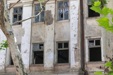 Kırık camları ve çatlak duvarları olan bir bina. Şehirde ağaçlarla çevrili. Eski beyaz beton bir yapı da dahil..