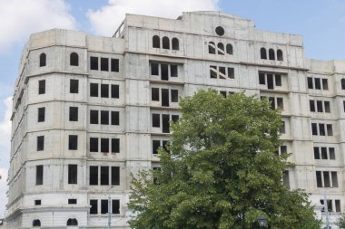 Gümüş taştan bir cephesi olan tamamlanmamış bir bina yuvarlak köşeli uzun pencereler ve tepesinde büyüyen büyük ağaçlar..