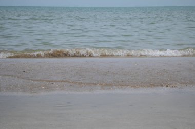 Açık gökyüzünün altında kumlu sahilde hafif bir dalga kopuyor..