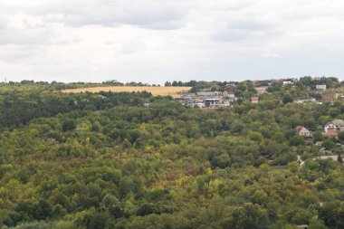 Scenic countryside landscape with green forest hills, rural houses and golden wheat field under cloudy summer sky in Moldova.