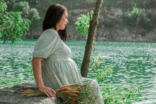 stock image Pregnant girl on the river bank. Waiting for the birth of a child. Woman with a bouquet of flowers made of reeds. A village girl sits by the water.