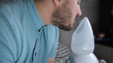 Yetişkin bir adam buhar soluyor, evde nebulizatörle hava yollarını tedavi ediyor. Adam solunum cihazıyla oturuyor, bronşit tedavisi, kovid, nebulizer, yakışıklı bir adam. Önleme ve tedavi