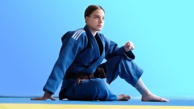 Kahverengi kemerli bir judo kızı. Genç bir judoka dövüşçüsü arka planda mavi kimonoyla poz veriyor. Japon dövüş sanatı. Dövüş sanatları eğitimi. Sağlıklı yaşam tarzı, spor anlayışı
