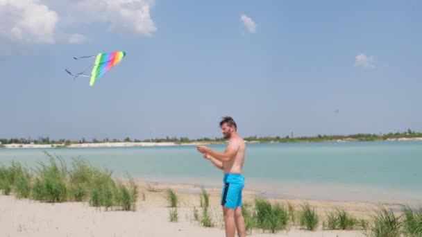 ビーチのスレンダーマンは 多色のカイトを飛んでいる 川岸でのアクティブなレクリエーション 大人のためのエンターテイメント — ストック動画