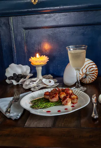 Candied bacon wrapped sea scallops over a bed of asparagus on fine China with a glass of white wine and a nautical table setting.