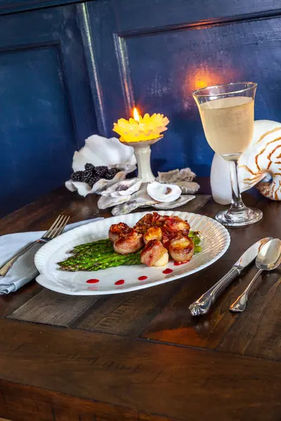 Candied bacon wrapped sea scallops over a bed of asparagus on fine China with a glass of white wine and a nautical table setting.
