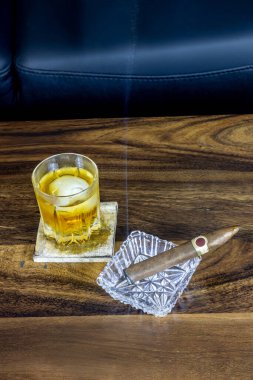 Crystal glass of whisky with an ice cube shaped like a skull inside and a fine cigar in a crystal ash tray on a walnut wood tabletop. clipart