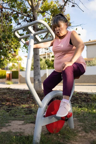 mature gray-haired woman with obesity exercising on an exercise bike in an outdoor training park.she is burning fat