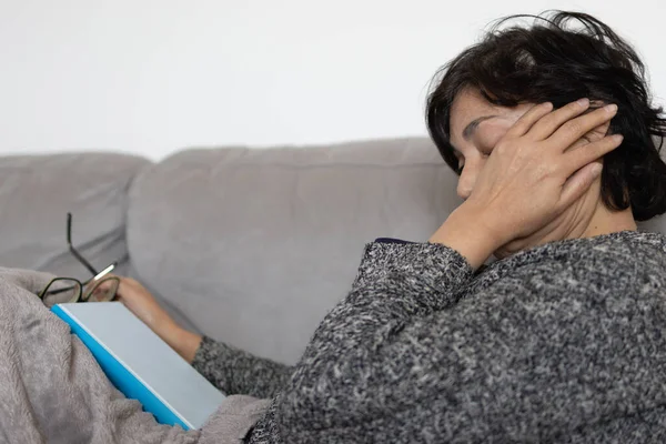 unrecognizable mature Middle Eastern brunette woman crying sadly sick from depression and loneliness. she is on the couch covered with a blanket. concept of reading self-help and psychology books.