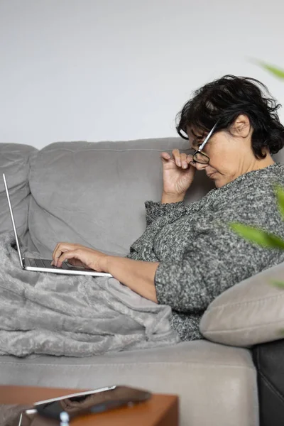 mature woman of Middle Eastern ethnicity, brunette, is covered with a warm blanket on the sofa. she is using her laptop computer concentrating on her digital online business.