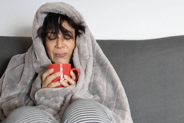 mature woman of Middle Eastern ethnicity enjoys drinking a cup of hot coffee in the early morning while covered with a warm blanket on the sofa at home. she is freshly awakened and sleepy.