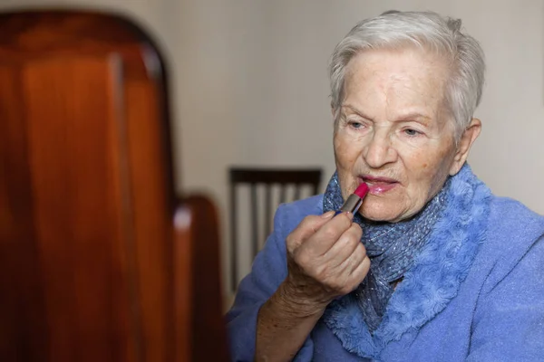 elderly woman with gray hair paints her lips red with a lipstick in front of the dressing table mirror. concept of makeup on elderly people