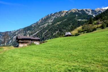 On a steep alpine meadow stands an abandoned very old farmhouse with a small church in the background. clipart