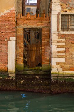 Venedik kanal binası yıpranmış tuğla, paslı metal bir kapı ve su yansımalarını gösteriyor. Kanal kenarındaki bozulma.